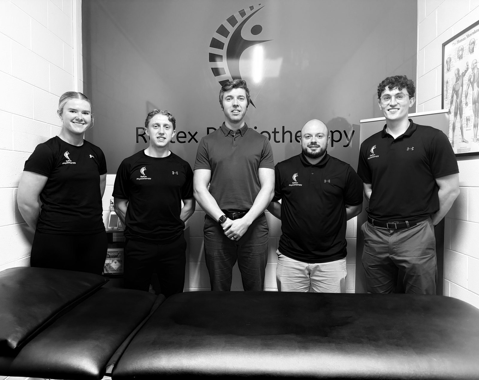 The five members of the Reflex Physiotherapy team standing in front of the Reflex logo wall