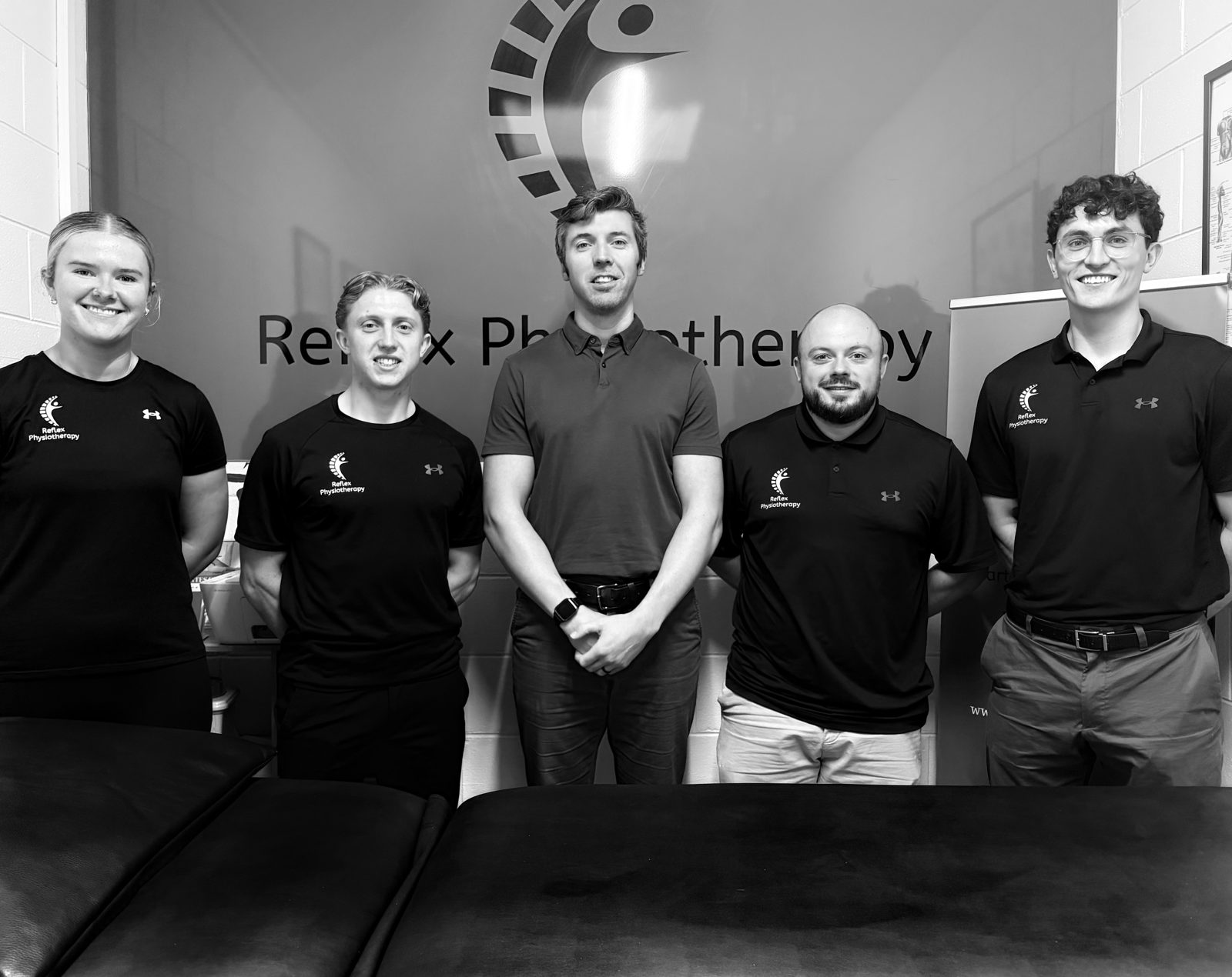 The five members of the Reflex Physiotherapy team standing in front of the Reflex logo wall