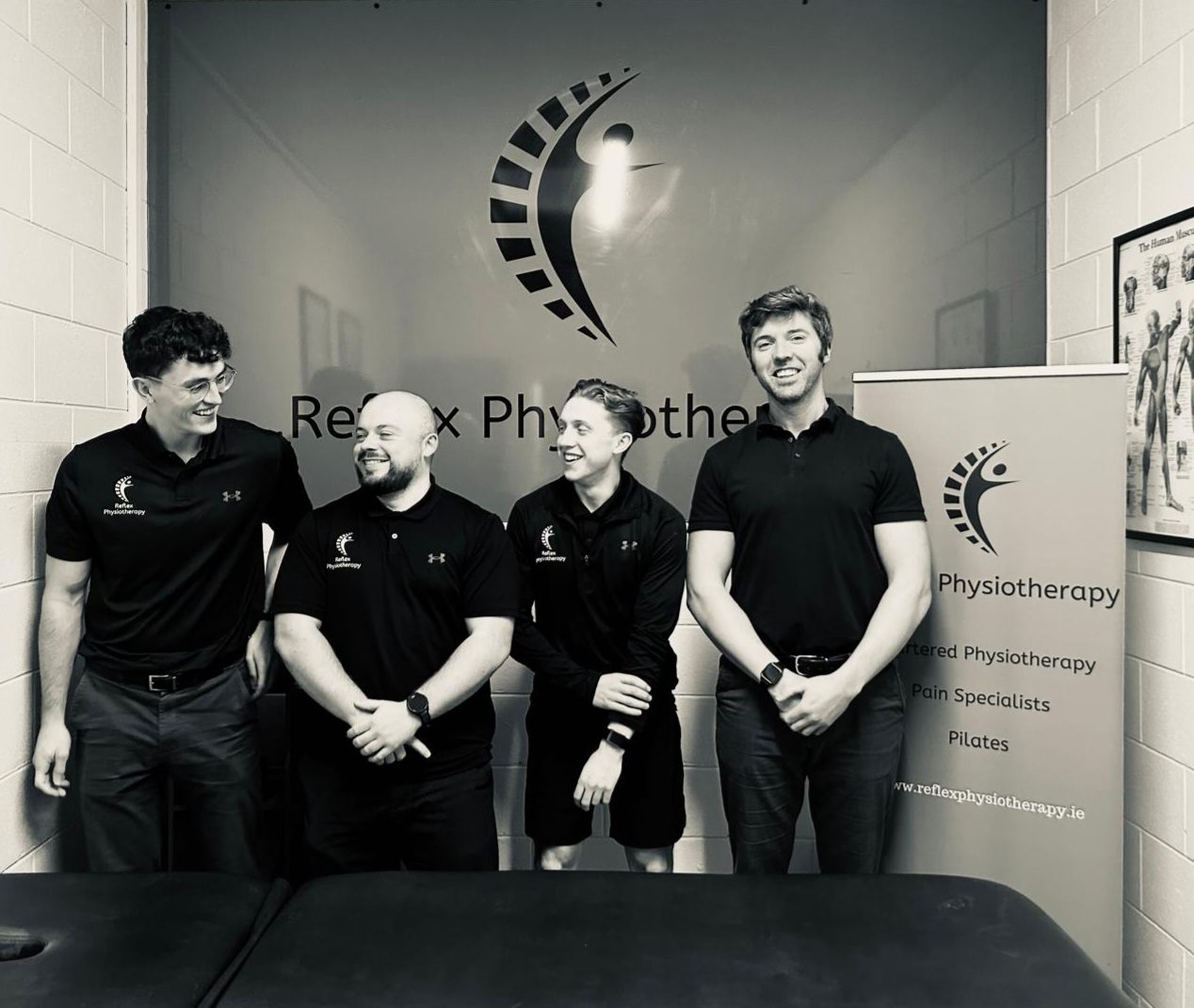 The Reflex Physiotherapy team in black Reflex polo shirts, standing in front of the Reflex logo wall